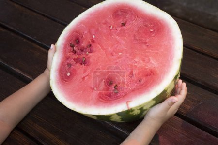 Photo for Kids hand holding half of watermelon on wooden table. Top view and copy space. - Royalty Free Image
