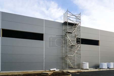 Fachada de panel sándwich gris de un edificio de almacén inacabado, andamios tubulares multinivel, fondo de cielo azul