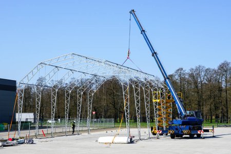 Photo for Assembly of the metal frame of the tent storage hangar with the help of a telescopic boom crane and a self propelled scissor lift - Royalty Free Image