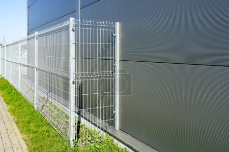 Téléchargez les photos : Une nouvelle clôture métallique galvanisée autour d'une nouvelle installation industrielle grise, de l'herbe verte et une voie pavée - en image libre de droit