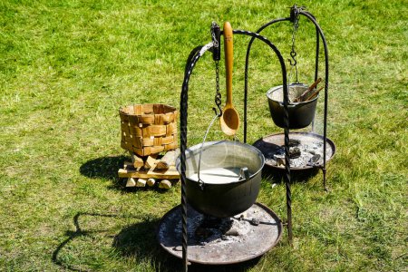 Cocinar en un fuego en un prado verde utilizando un soporte de metal con una olla colgante y una base de metal para leña, marco de cocción