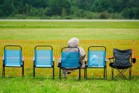 Photo for Folding chairs with a lone spectator at the edge of a green field waiting for the show to start, orange plastic temporary portable fence, rear view - Royalty Free Image