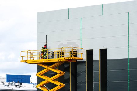Foto de Gran plataforma elevadora de tijera autopropulsada con trabajadores en dos tonos gris nuevo edificio industrial paneles sándwich pared, elevador industrial en acción - Imagen libre de derechos