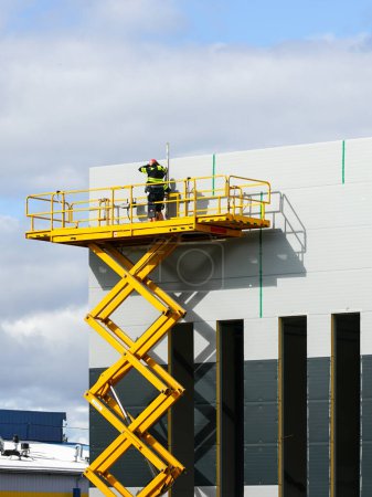 Foto de Amplia plataforma de trabajo de elevación de tijera aérea elevada amarilla con trabajadores en la pared de paneles sándwich de edificio industrial nuevo gris, elevador industrial - Imagen libre de derechos