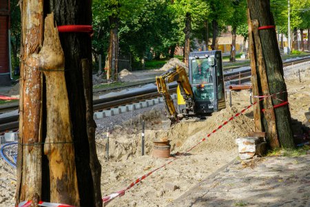 Téléchargez les photos : Vue de reconstruction de la rue et de la voie de tramway, protection des troncs d'arbres contre les dommages avec couvertures de panneaux, excavatrice creuse une tranchée - en image libre de droit
