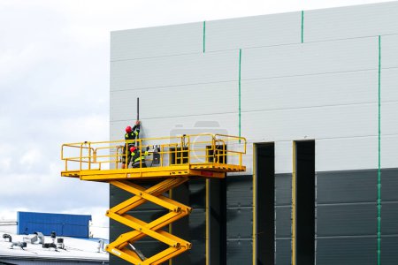 Foto de Amplia plataforma de trabajo de elevación de tijera aérea elevada amarilla con trabajadores en la pared de paneles sándwich de edificio industrial nuevo gris, elevador industrial - Imagen libre de derechos