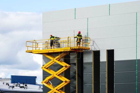 Téléchargez les photos : Grande plate-forme de travail élévatrice à ciseaux surélevée jaune avec travailleurs au mur gris de nouveaux panneaux sandwich de bâtiment industriel, élévateur industriel - en image libre de droit