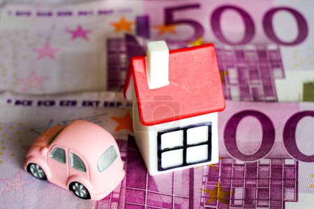 Small white toy house with red roof and small pink car on 500 euro banknote background, house and car price concept