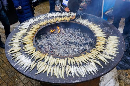 Many grilled smelt fish on a large round flat grill with open fire hole in the center, outdoor seafood cooking, frying fish on an open fire grill