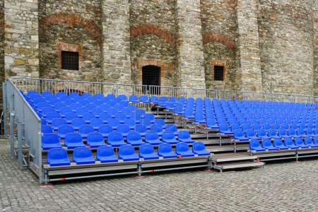 Sala de conciertos al aire libre con filas de asientos azules vacíos, estructura metálica móvil con asientos de plástico azul en el patio de un castillo histórico