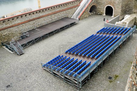 Salle de concert extérieure avec scène et rangées vides de sièges d'audience, structure métallique mobile avec des sièges en plastique bleu dans la cour d'un château historique