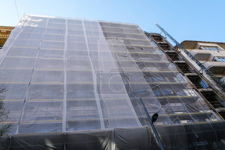 Graues Sicherheitsnetz um eine große Stadthausfassade während Umbau und Sanierung, Gerüst mit grauem Schutzanstrich, provisorischer Aufzug