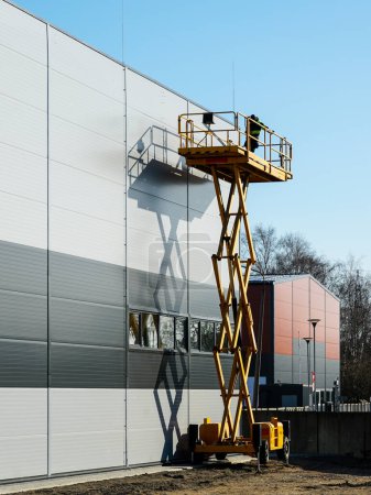 Foto de Plataforma de trabajo de elevación de tijera autopropulsada amarilla elevada con trabajador para inspección de paredes de edificios industriales, fondo azul del cielo - Imagen libre de derechos