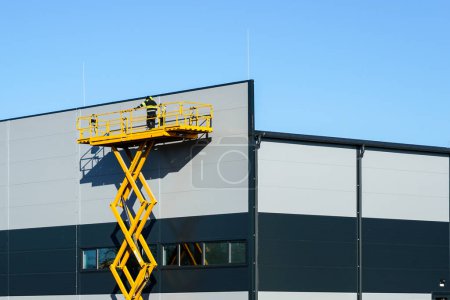 Photo for High elevated yellow self propelled scissor lift working platform with worker for industrial building wall inspection, blue sky background - Royalty Free Image