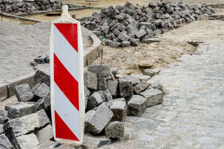 Street paving with historical hammered granite paving stones, city street surface restoration, cubes rocks, red and white striped vertical warning sign
