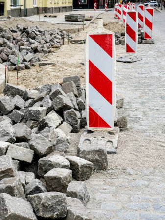 Pavimentación de calles con adoquines históricos de granito martillado, restauración de la superficie de la calle de la ciudad, rocas de cubos, señal de advertencia vertical a rayas rojas y blancas