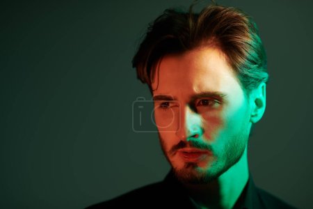Portrait of a handsome serious brunet man in a black shirt looking  intensely away. Mixed-coloured light on a dark background with copy space. Men's beauty, emotions.