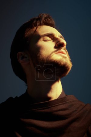 A portrait of a masculine brunet man in a black cardigan who stands with his eyes closed and retreats into himself, turning his head towards the setting sun against a dark blue sky. Emotions.