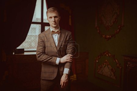 Respectable lifestyle. Portrait of a well-groomed young man in an elegant plaid suit and bow tie adjusting his sleeve. Luxurious vintage interior. Men's fashion.