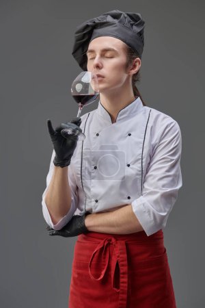 Porträt eines professionellen anspruchsvollen jungen Kochs in gepflegter Uniform, der vorzüglichen Rotwein riecht.