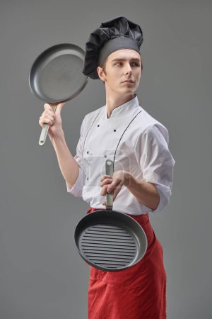 Young handsome chef in neat  uniform poses with frying pans in his hands. Gray studio background. Professional kitchen utensils.