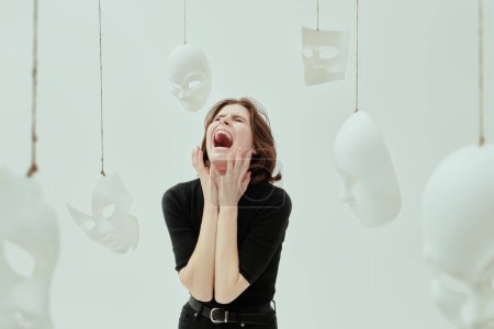 A girl in black clothes screams furiously, surrounded by white masks. Human roles. Hypocrisy. Mental disorders. Studio photo on a white background.