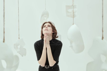 Theater of life. The unfortunate girl stands with her eyes raised to the sky and hands clasped in prayer, surrounded by masks. Hypocrisy. Studio photo on a white background.