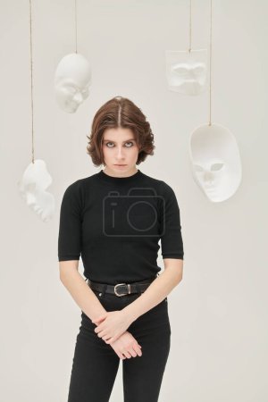 Theater of life. A girl in black clothes stands in a white room surrounded by various masks looking intently at the camera. Hypocrisy. Human roles. Mental disorders.