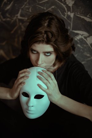 Trastornos mentales. Una chica vestida de negro se sienta apoyada en una pared grunge apretando un papel de máscara en sus manos y mirando nerviosamente hacia arriba. Papeles humanos. Hipocresía. 
