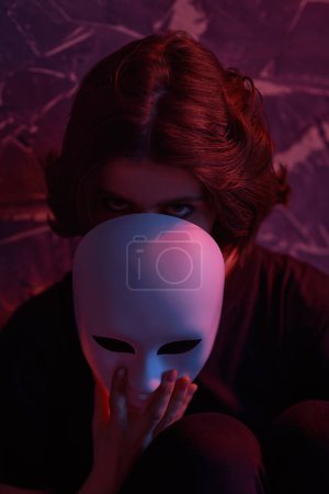 Hipocresía. Una chica vestida de negro mira a la cámara desde detrás de una máscara contra una pared grunge oscura con luz roja. Papeles humanos. Trastornos mentales. 
