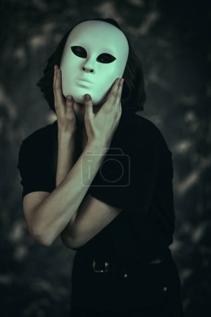 Une fille en vêtements noirs essaie un rôle de masque, debout sur un fond sombre grunge. Les rôles humains. Hypocrisie. Troubles mentaux.