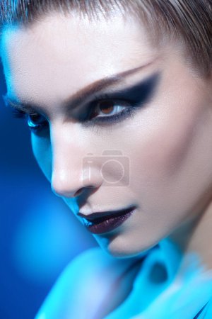 Gros plan portrait d'une fille mannequin au maquillage sombre et agressif détournant les yeux. Studio tourné sur fond bleu. Maquillage et cosmétiques. 