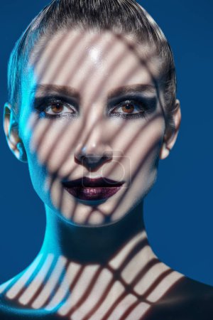 Portrait d'une fille mannequin avec un maquillage sombre posant avec des ombres sur son visage. Studio tourné sur fond bleu. Maquillage et cosmétiques. 