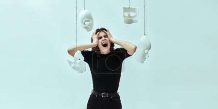 Una chica vestida de negro grita desesperada sosteniendo su cabeza, rodeada de máscaras blancas. Papeles humanos. Hipocresía. Trastornos mentales. Foto del estudio sobre fondo blanco.