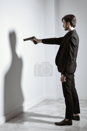 Portrait d'un brunettiste vêtu d'un costume noir, debout devant le mur, pointant un fusil à la main vers son ombre. Se battre avec soi-même. L'ennemi est à l'intérieur.