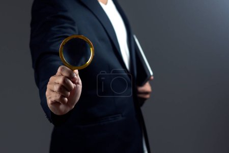 Businessman is using magnifying glass and hold digital tablet against dark background.