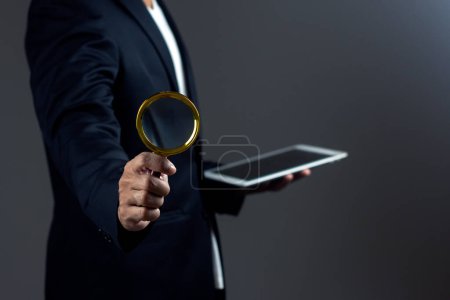 Businessman is using magnifying glass and using digital tablet against dark background.