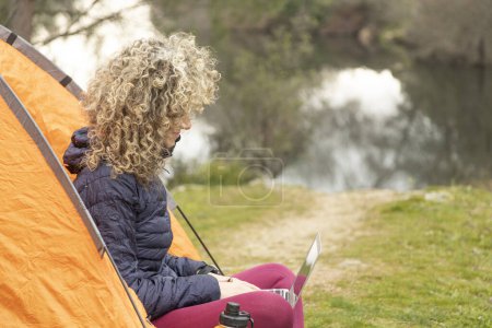 Photo for Curly and blonde hair, mountaineer woman camps with his orange tent, teleworking with his laptop - Royalty Free Image