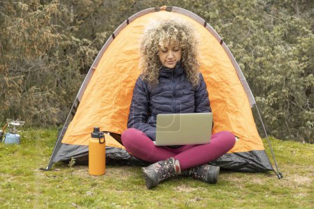 Téléchargez les photos : Cheveux bouclés et blonds, camps de montagne femme avec sa tente orange, télétravail avec son ordinateur portable - en image libre de droit