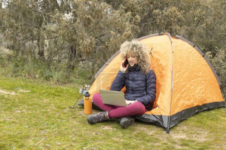 Photo for Curly and blonde hair, mountaineer woman camps with his orange tent, teleworking with his laptop - Royalty Free Image