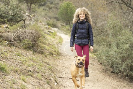 Photo for Curly and blonde hair, mountaineer woman and her dog walking through the mountains. - Royalty Free Image