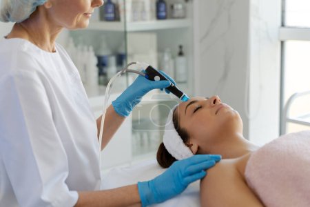 Photo for Cleansing procedure. Nice pretty woman lying with her eyes closed while having a cleansing procedure on her face. Senior doctor working with smile - Royalty Free Image