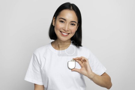 Foto de Mujer asiática positiva demostrando un desfibrilador cardioversor implantable (DAI) en las manos mientras mira a la cámara con una sonrisa agradable. Cuidado de la salud y concepto del corazón - Imagen libre de derechos