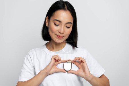 Foto de Mujer asiática morena buscando Desfibrilador cardioversor implantable (DAI) mientras lo sostiene a manos sobre fondo blanco. Cuidado de la salud y concepto del corazón - Imagen libre de derechos