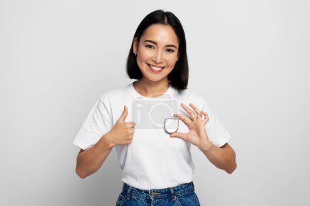 Foto de Mujer bastante asiática sosteniendo desfibrilador cardioversor implantable (DAI) y mostrando un gesto fresco en el fondo blanco. Cuidado de la salud y concepto del corazón - Imagen libre de derechos
