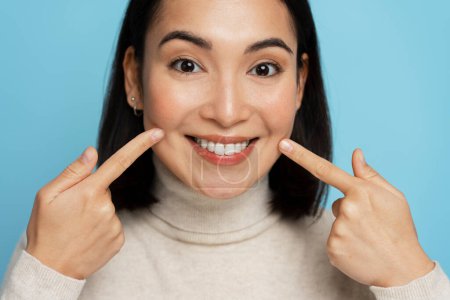 Photo for Portrait of pretty cheerful woman points index fingers at smile shows white teeth, looking at camera. Indoor studio shot isolated on blue background - Royalty Free Image