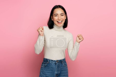 Yes. Portrait of happy cheering young woman standing with raised arms, screaming and celebrating her victory. Indoor studio shot, isolated on pink background 