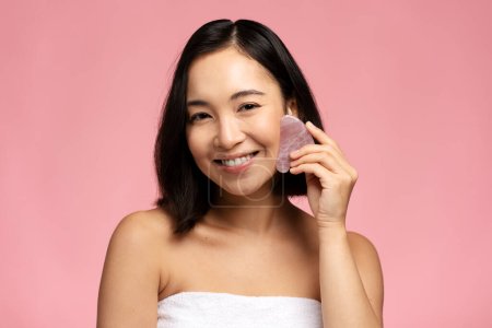 Portrait of smiling beautiful naked asian woman using facial gua sha jade board isolated on pink background. People skin care concept