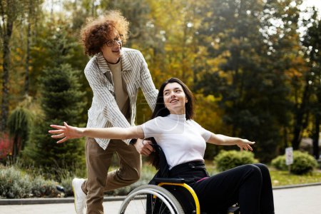 Téléchargez les photos : Cheerful, smiling guy is driving a wheelchair with a girl outdoors. A cheerful girl spread her arms to the sides, like the wings of an airplane, on the street. - en image libre de droit