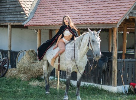 Foto de Sensual princesa morena con sexy bikinis blanco montando un caballo. Retrato de una chica con capa y corona y su caballo. Chica interactuando y divirtiéndose con un caballo en el estilo cuento de hadas historia - Imagen libre de derechos